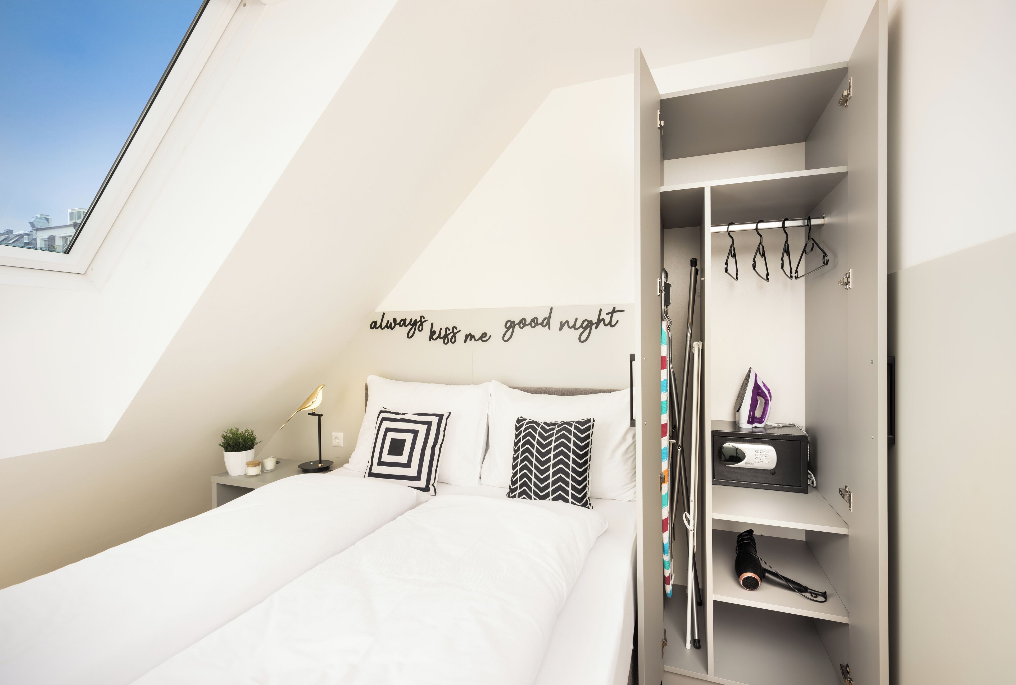 Een slaapkamer met dakraam, een bed met witte beddengoed, een nachtkastje met lamp en plant, en een kledingkast met kleren.