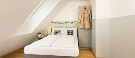 Quarto com teto inclinado, cama com roupa de cama branca, mesa de cabeceira com abajur, casaco pendurado na parede e uma pequena planta.