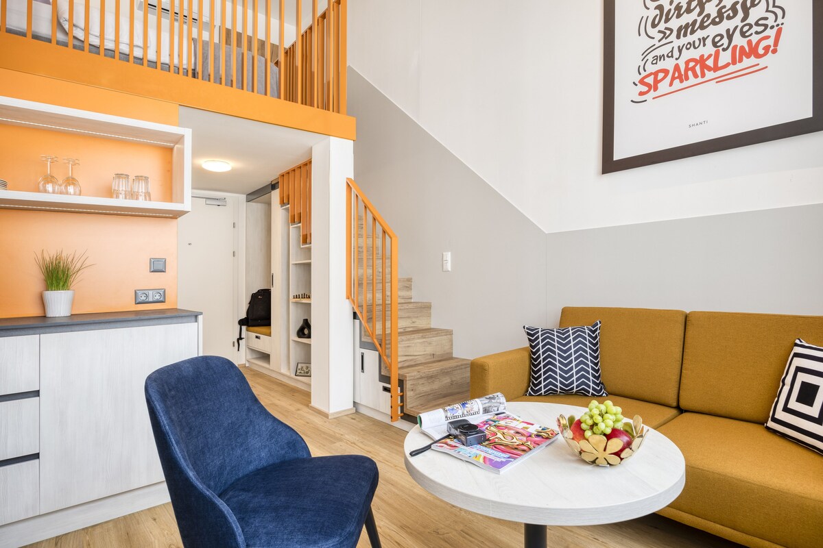 A modern living room with a sofa, a round coffee table with magazines and fruit, a blue chair, a kitchen area with a counter and shelves, and a staircase with orange railings.