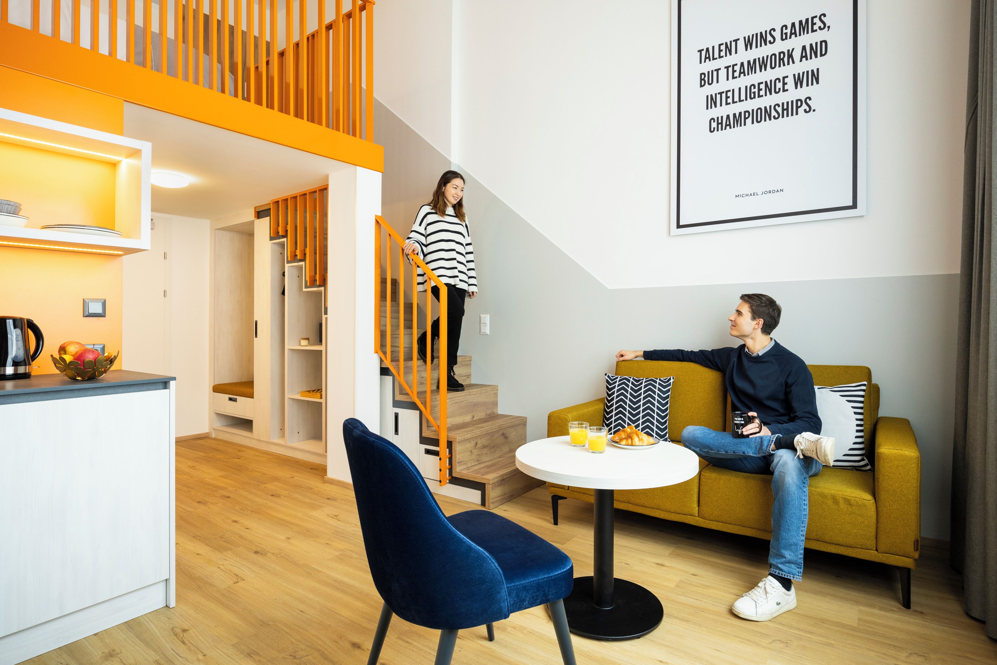 A modern interior with a staircase, a sofa, a dining area, and a wall with a motivational quote.