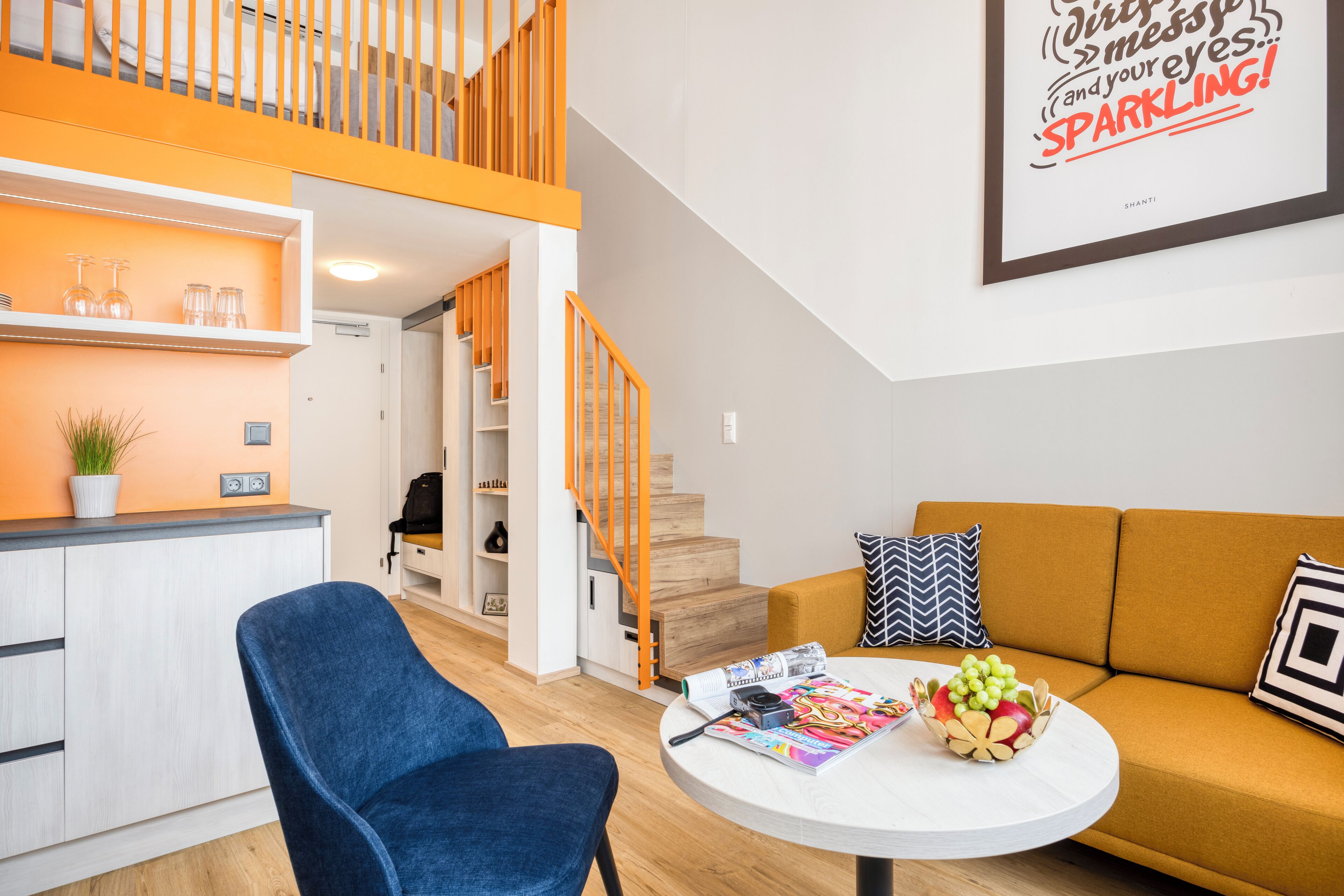Una sala de estar moderna con un sillón azul, una mesa redonda con revistas y una cámara, un sofá con almohadones estampados, y una escalera con barandillas naranjas.