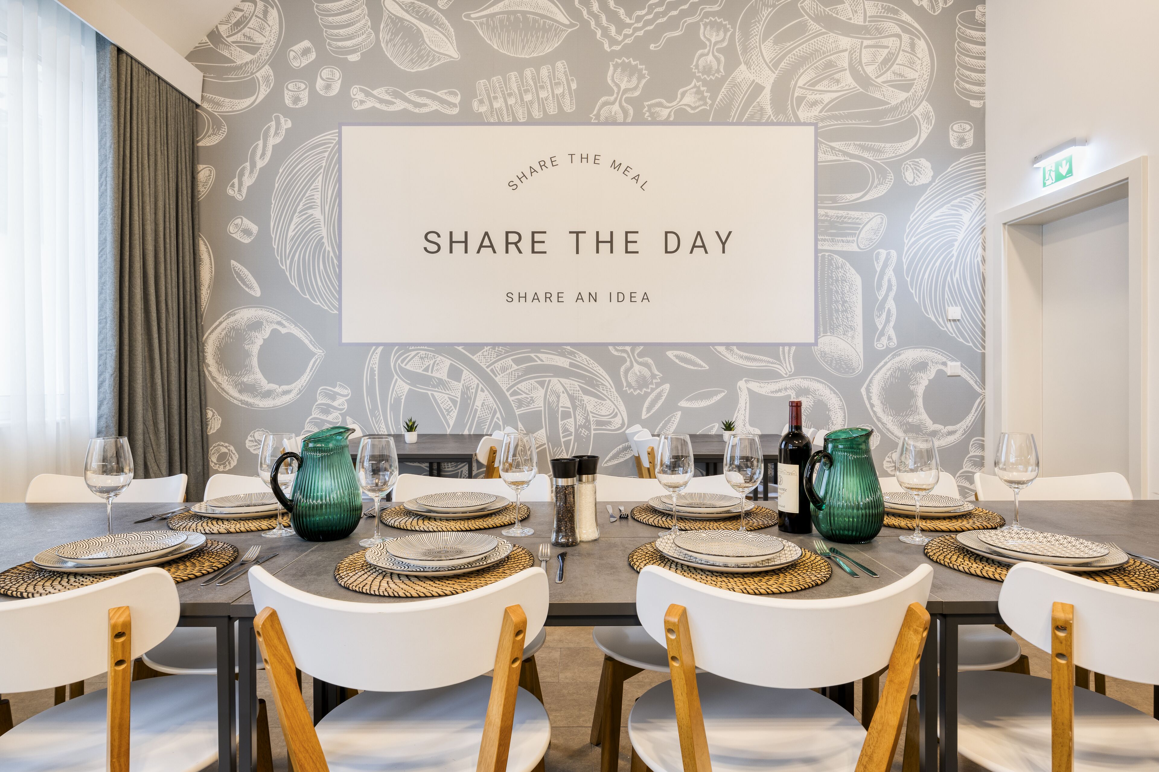 A dining area with a table set for six, featuring woven placemats, white chairs, and a decorative wall with a 'Share the Meal' sign.