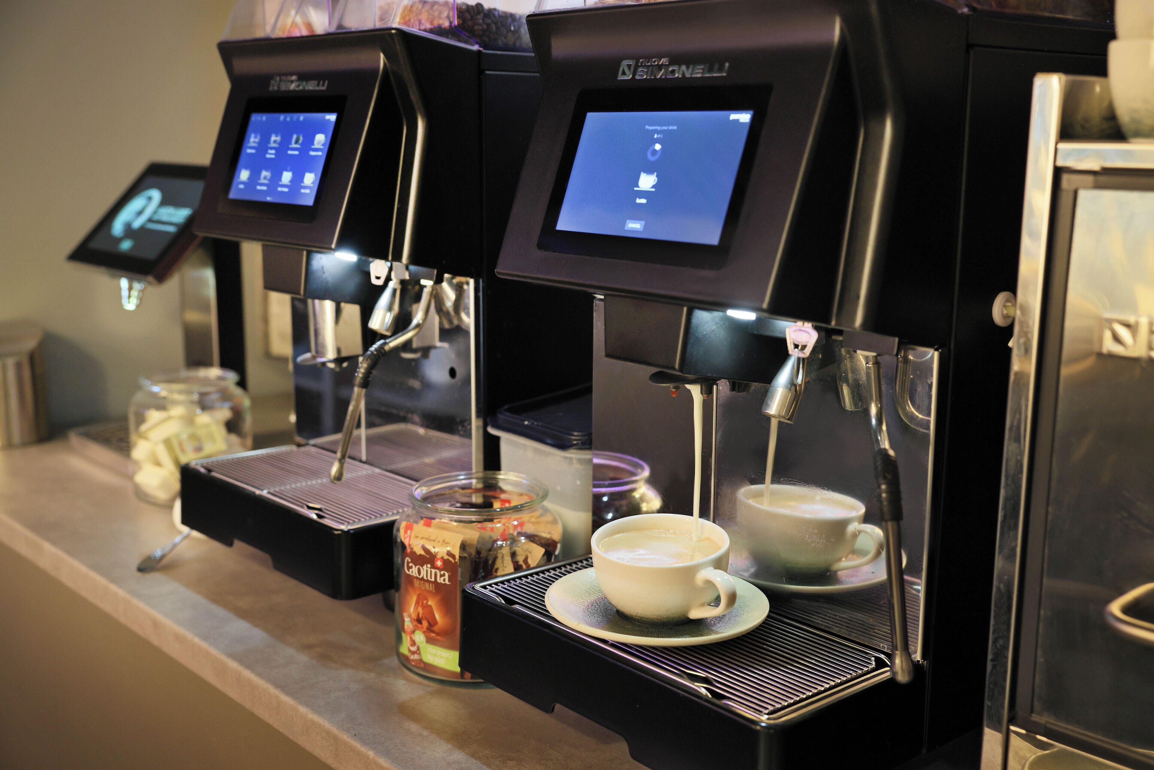 Una cafetera con pantalla digital, una taza de café y un tarro de leche condensada sobre una encimera.