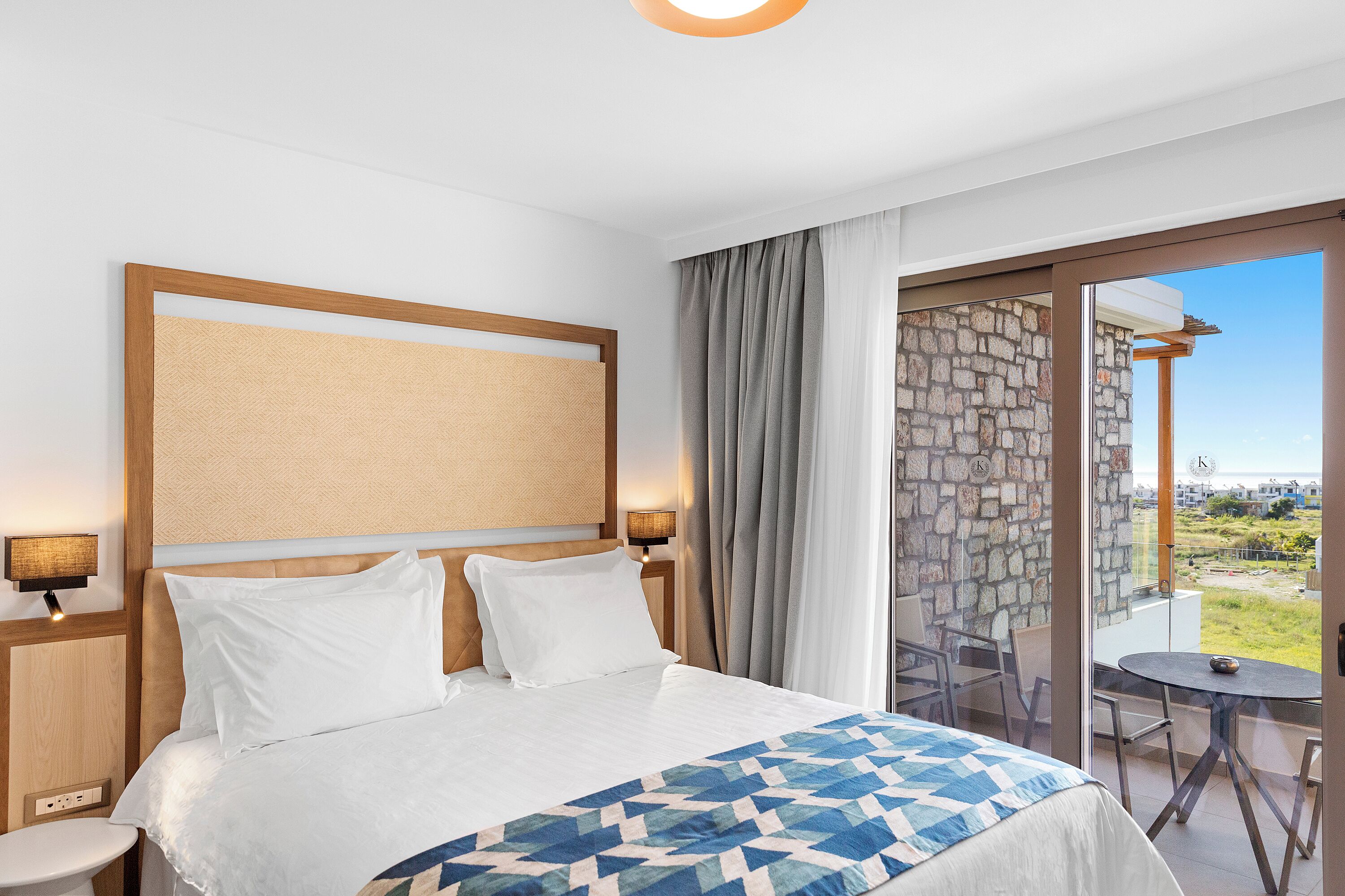 Hotellihuone, jossa on suuri sänky, kaksi yöpöytää, yölamppu, parveke, jossa on pöytä ja tuolit, sekä kiviseinä.