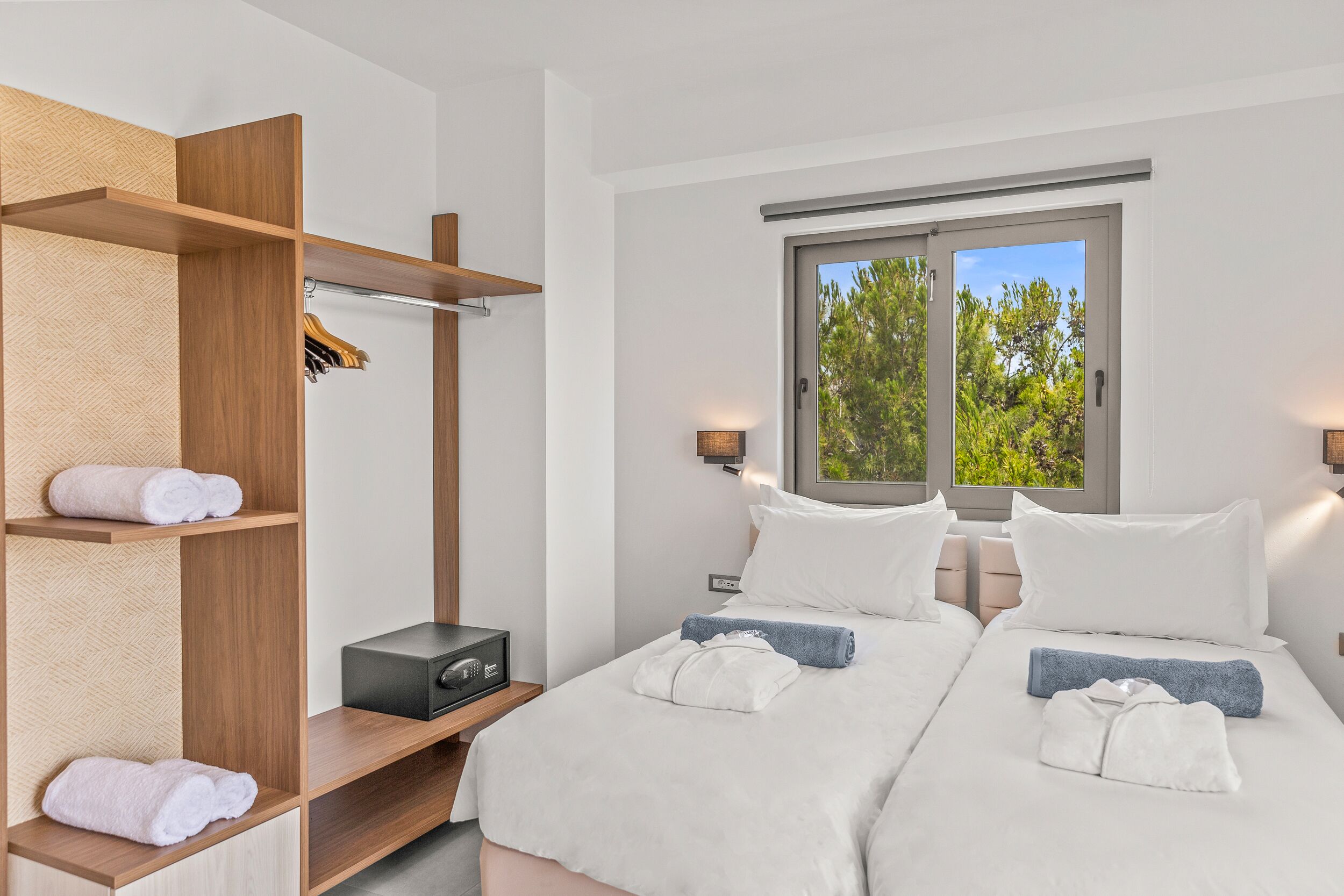 Ein Hotelzimmer mit zwei Betten, einem Holzschrank, einem Fenster mit Blick auf Bäume und einem Nachttisch mit Lampe.
