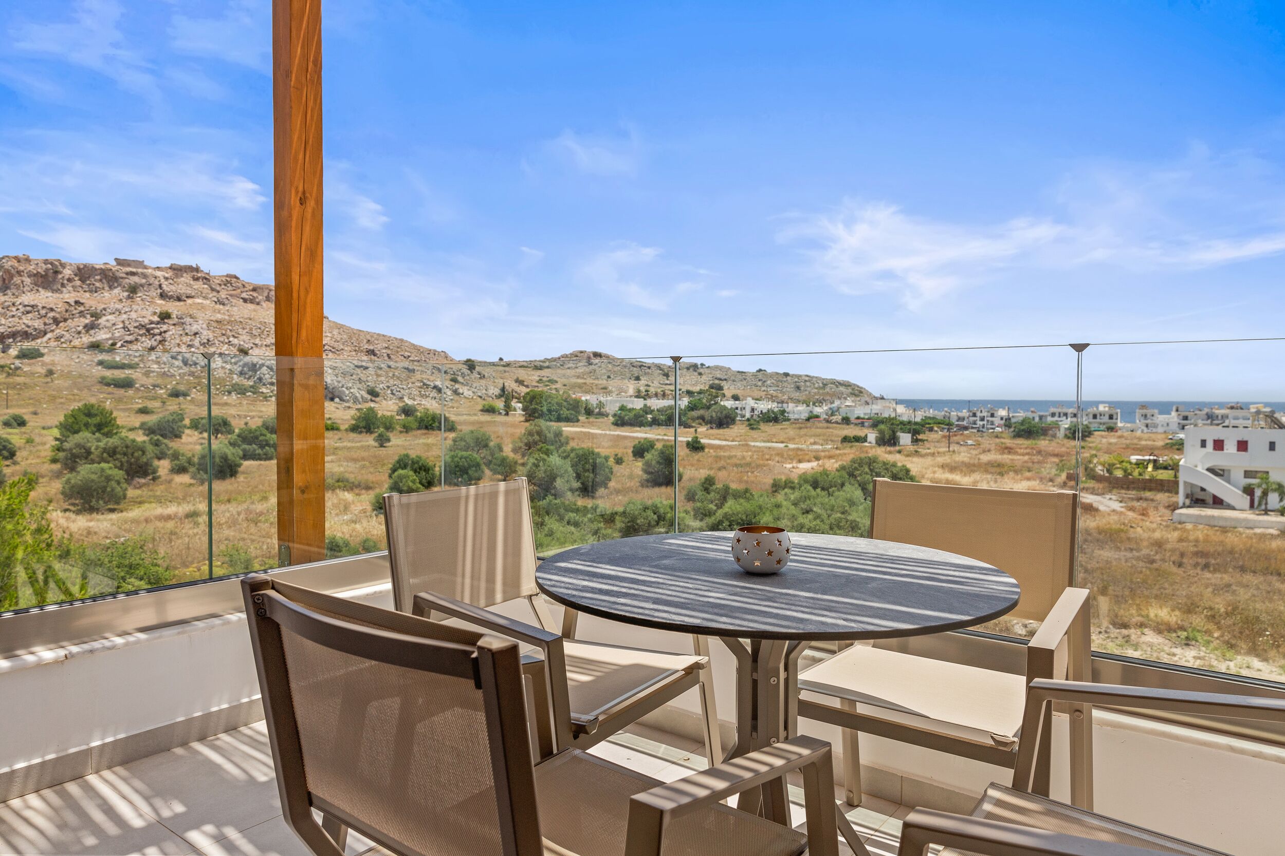 A balcony with a round table and chairs, overlooking a hilly landscape with sparse vegetation and a distant view of buildings.