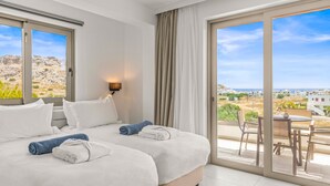 Habitación de hotel con dos camas, un balcón con mesa y sillas, y vistas al paisaje.