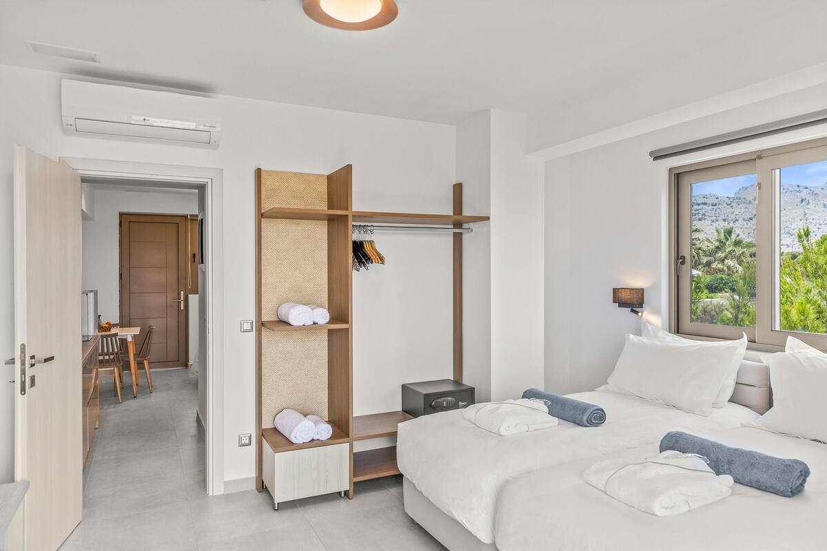 A modern hotel room with a large bed, a built-in wardrobe, a bedside table, and a view of greenery through the window.