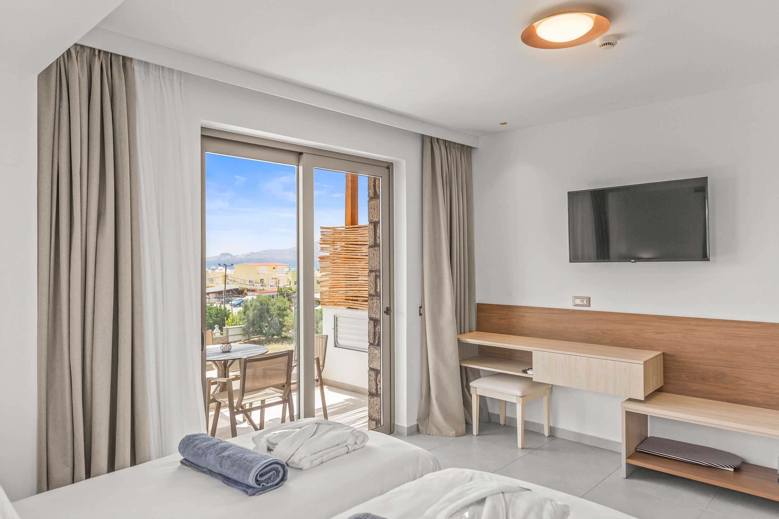 Ett hotellrum med en säng, ett skrivbord, en TV och en balkong med utsikt över staden.
