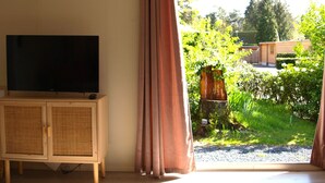 A room with a television on a wooden cabinet, a large window with curtains, and a view of a garden with greenery and a wooden structure.
