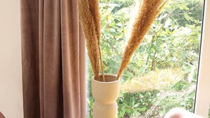A window with a view of greenery, a vase with pampas grass, and a table with small containers.