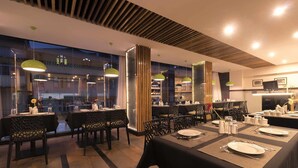 Un restaurant avec des tables dressées pour le service, un bar et une vue sur le paysage urbain grâce à de grandes fenêtres.