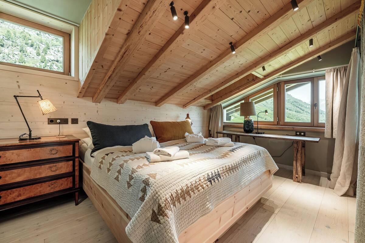 A bedroom with a wooden ceiling, a bed with a patterned quilt, a nightstand with a lamp, and a window with a view of hills.