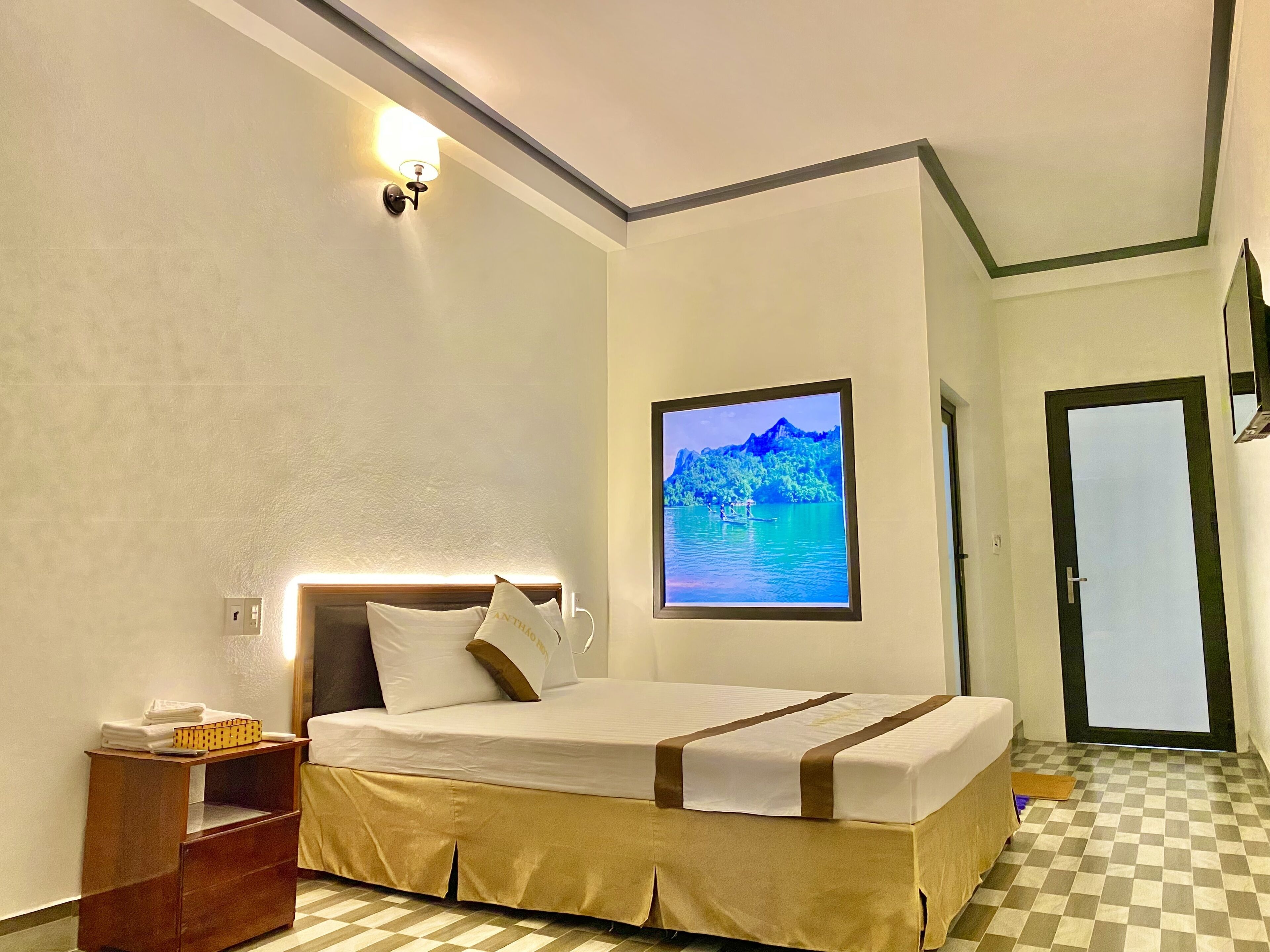A hotel room with a bed, a nightstand, a wall-mounted lamp, a framed picture, and a checkered floor.