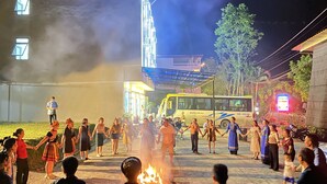 Eine Gruppe von Menschen hält Händchen im Kreis um ein Lagerfeuer, im Hintergrund ein Bus und ein Gebäude.