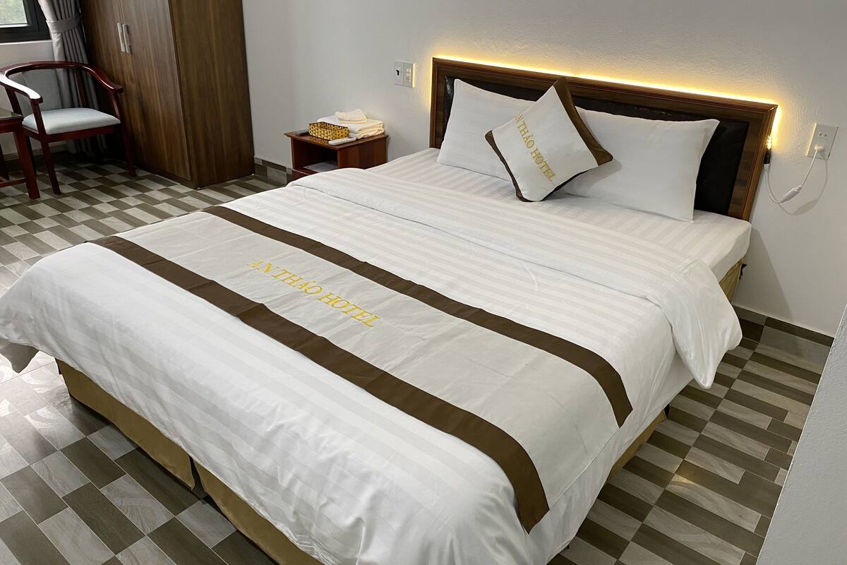 Quarto de hotel com cama bem arrumada, mesa de cabeceira com abajur e uma cadeira.