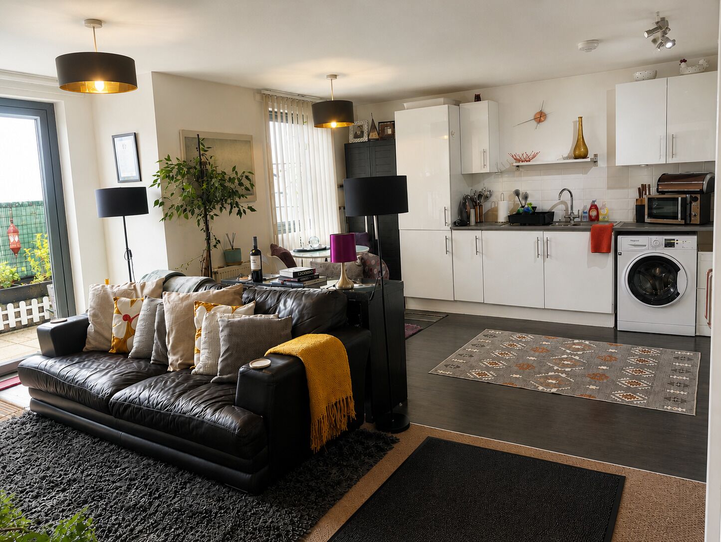 Una sala moderna con un sofá de cuero negro, una mesa de centro y un televisor. Junto a ella, una cocina con armarios blancos, una lavadora y una zona de comedor con mesa y sillas.