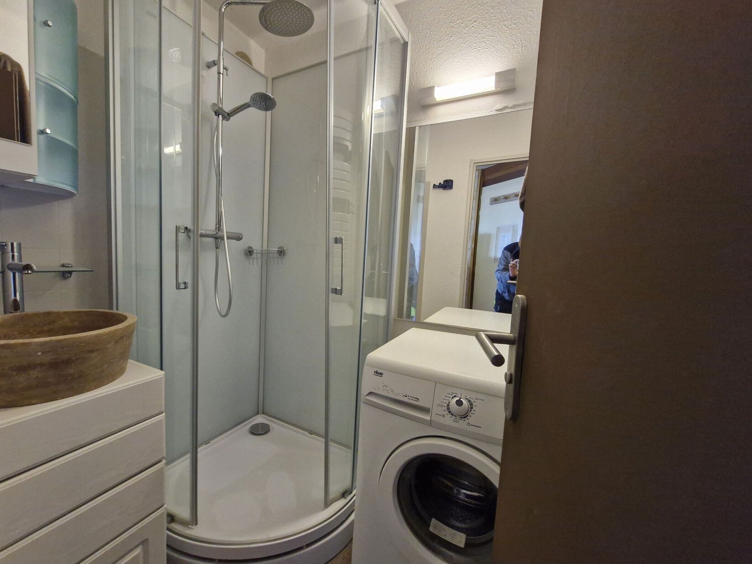 A compact bathroom with a glass-enclosed shower, a wooden basin, and a front-loading washing machine.