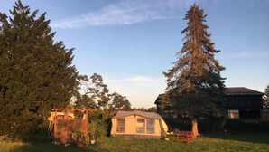 Una piccola casa a un piano con tetto spiovente, circondata da un prato verde e alti alberi sempreverdi.