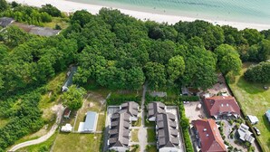An aerial view of a residential area with a beach, dense greenery, and several houses with red roofs. - Appartementanlage Eldena Wohnung Nr. 10 (Mönchgut OT Lobbe)