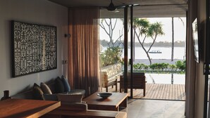 Un moderno salón con una mesa de centro de madera, un sofá y un gran cuadro abstracto en la pared. Se ve una masa de agua y palmeras a través del ventanal.