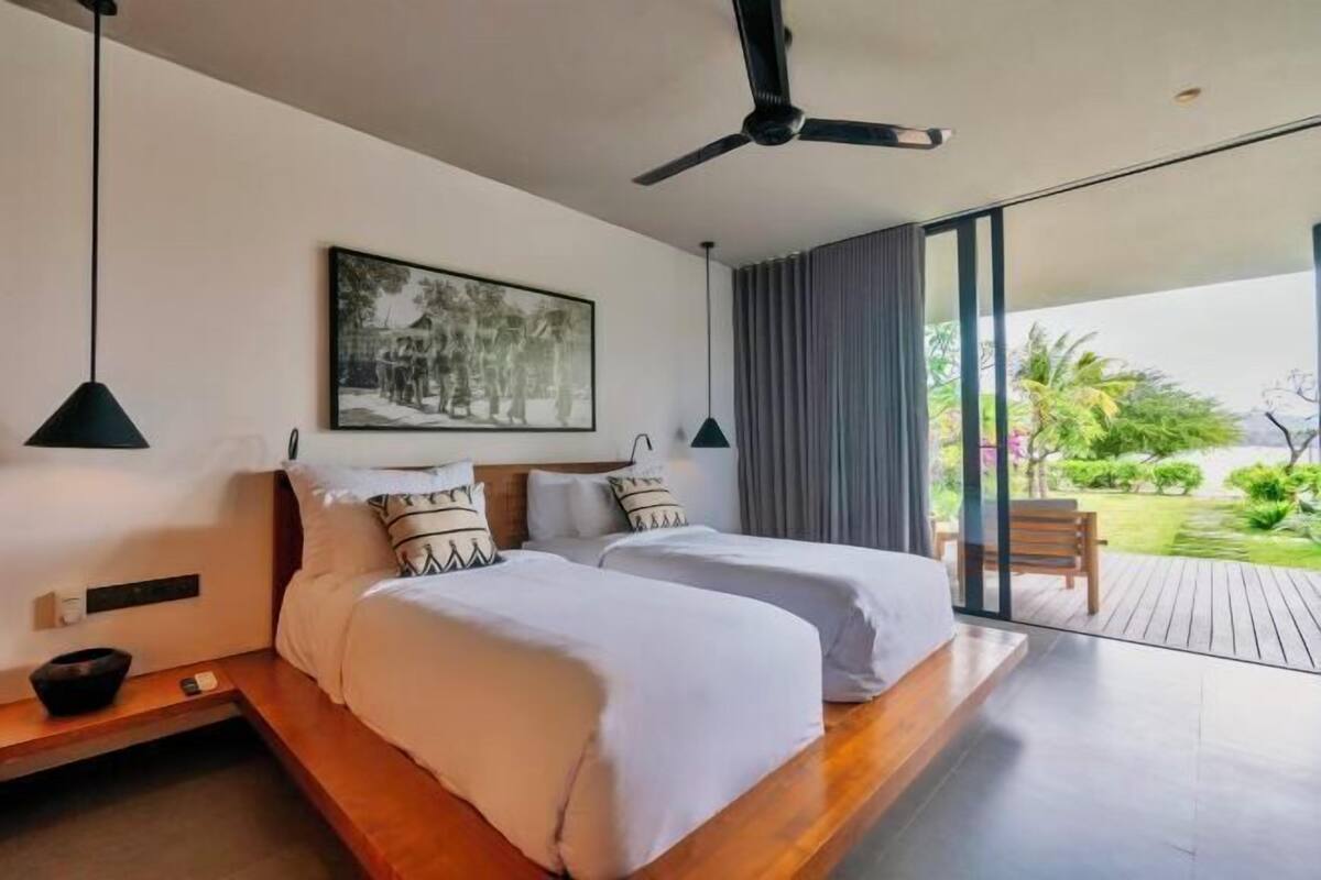 A modern hotel room with two beds, a ceiling fan, a large window, and a view of the outdoors.