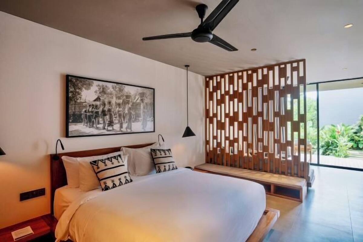 A modern hotel room with a large bed, a ceiling fan, a wooden partition, and a large window.