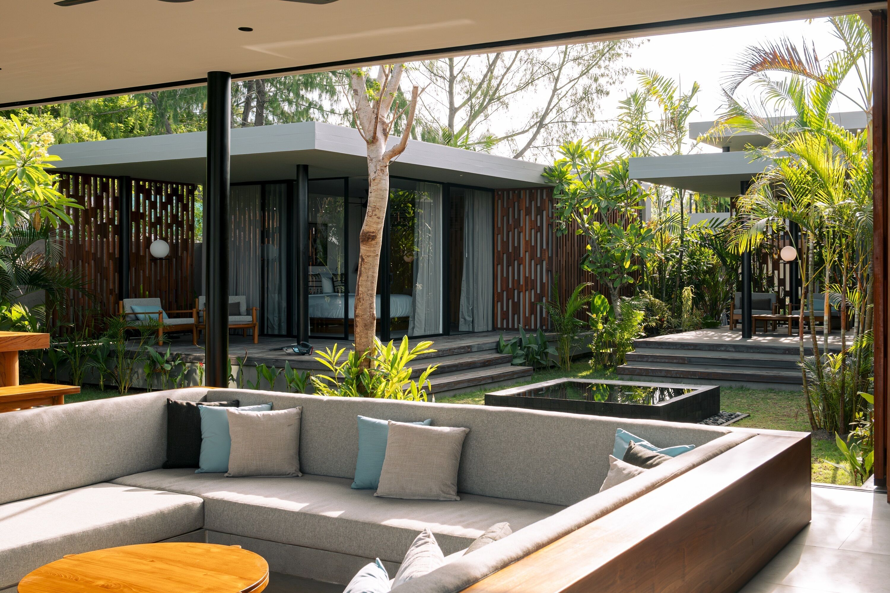 A modern outdoor living area with a sofa, a round wooden table, and a view of a house with large windows and a garden.