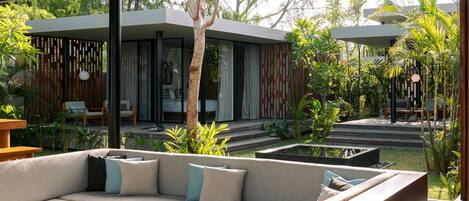 A modern outdoor living area with a sofa, a round wooden table, and a view of a house with large windows and a garden.
