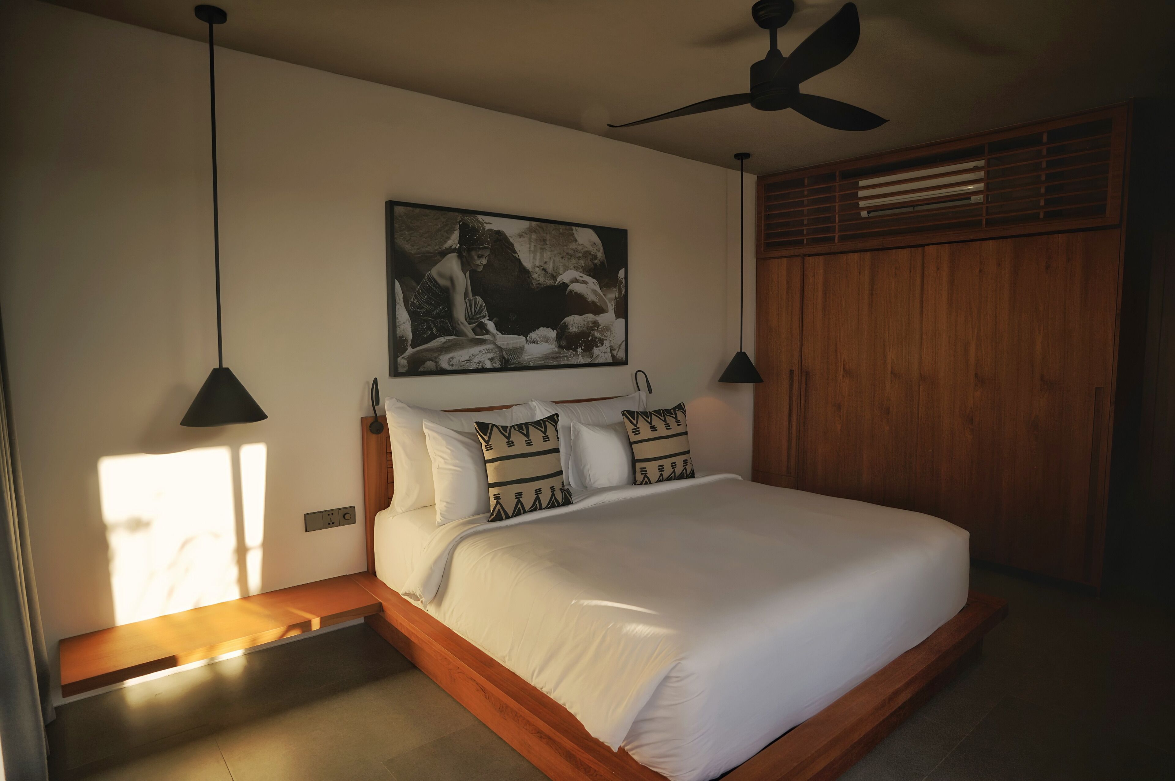 Un dormitorio con cama, ventilador de techo, cabecera de madera, un cuadro en la pared y una ventana con luz solar.