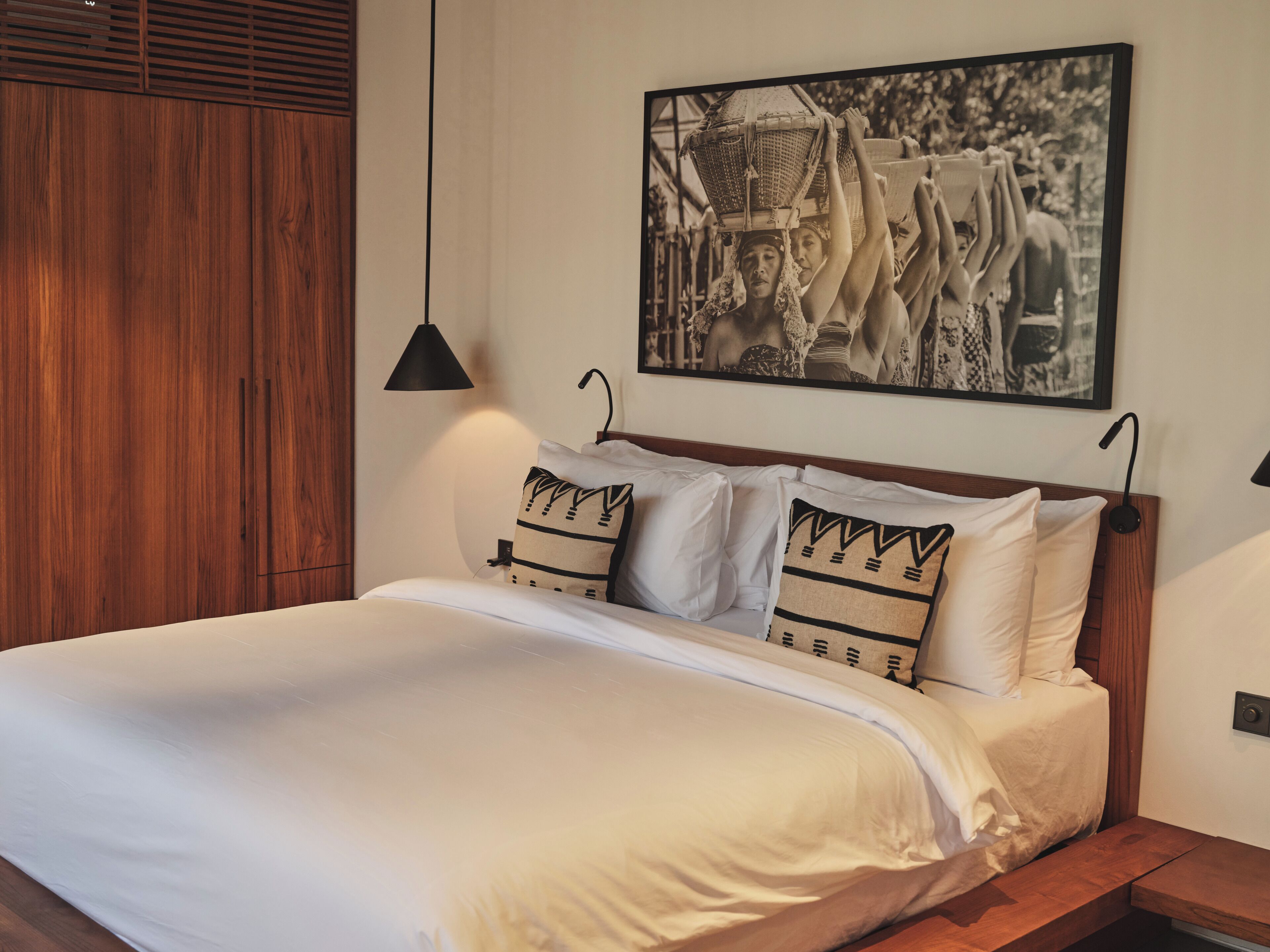 A neatly made bed with white linens and patterned pillows, a wooden headboard, and a black pendant light.