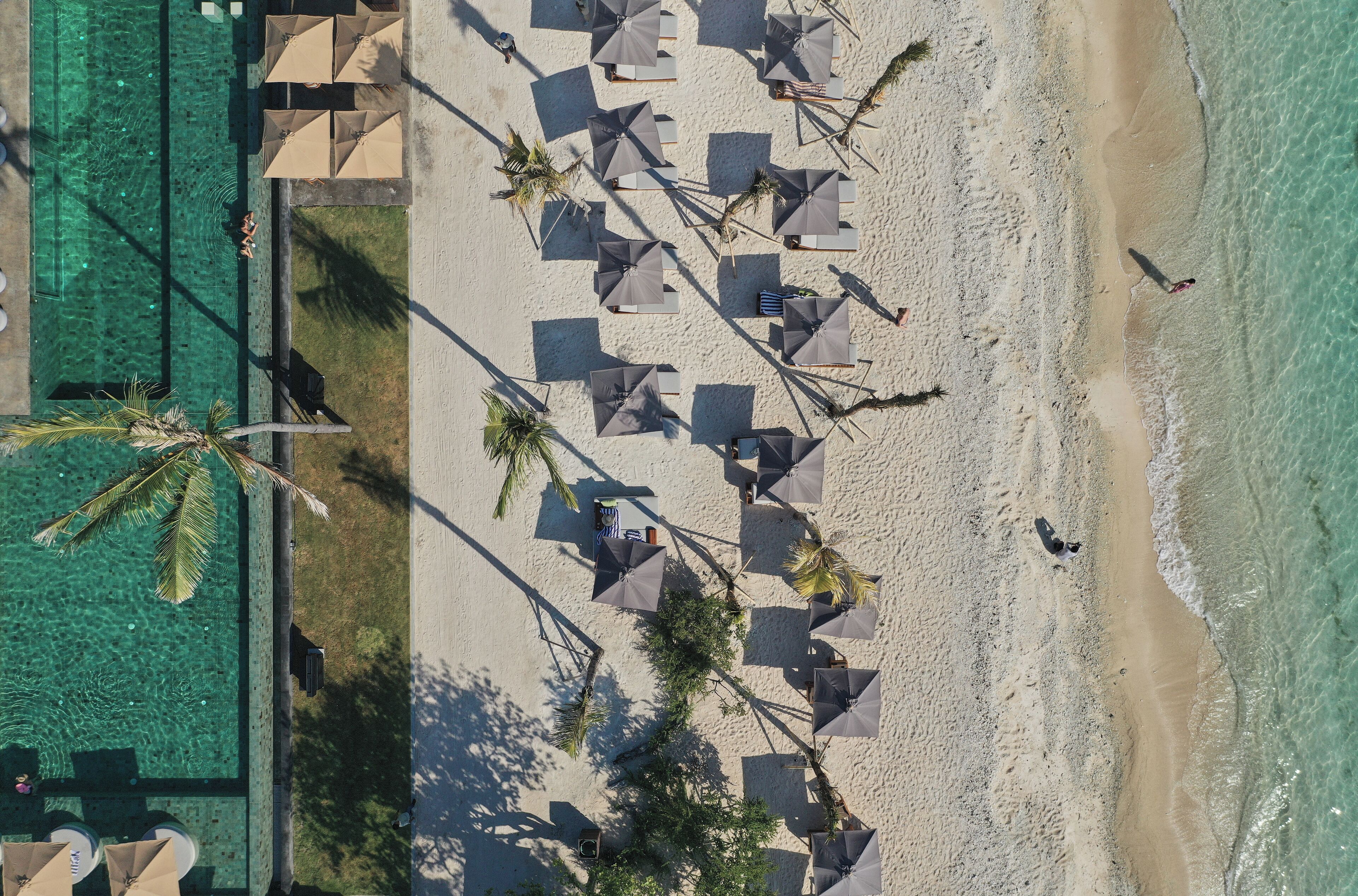 Luftaufnahme eines Strandes mit Sonnenschirmen, Liegestühlen, einem Schwimmbad und Palmen.