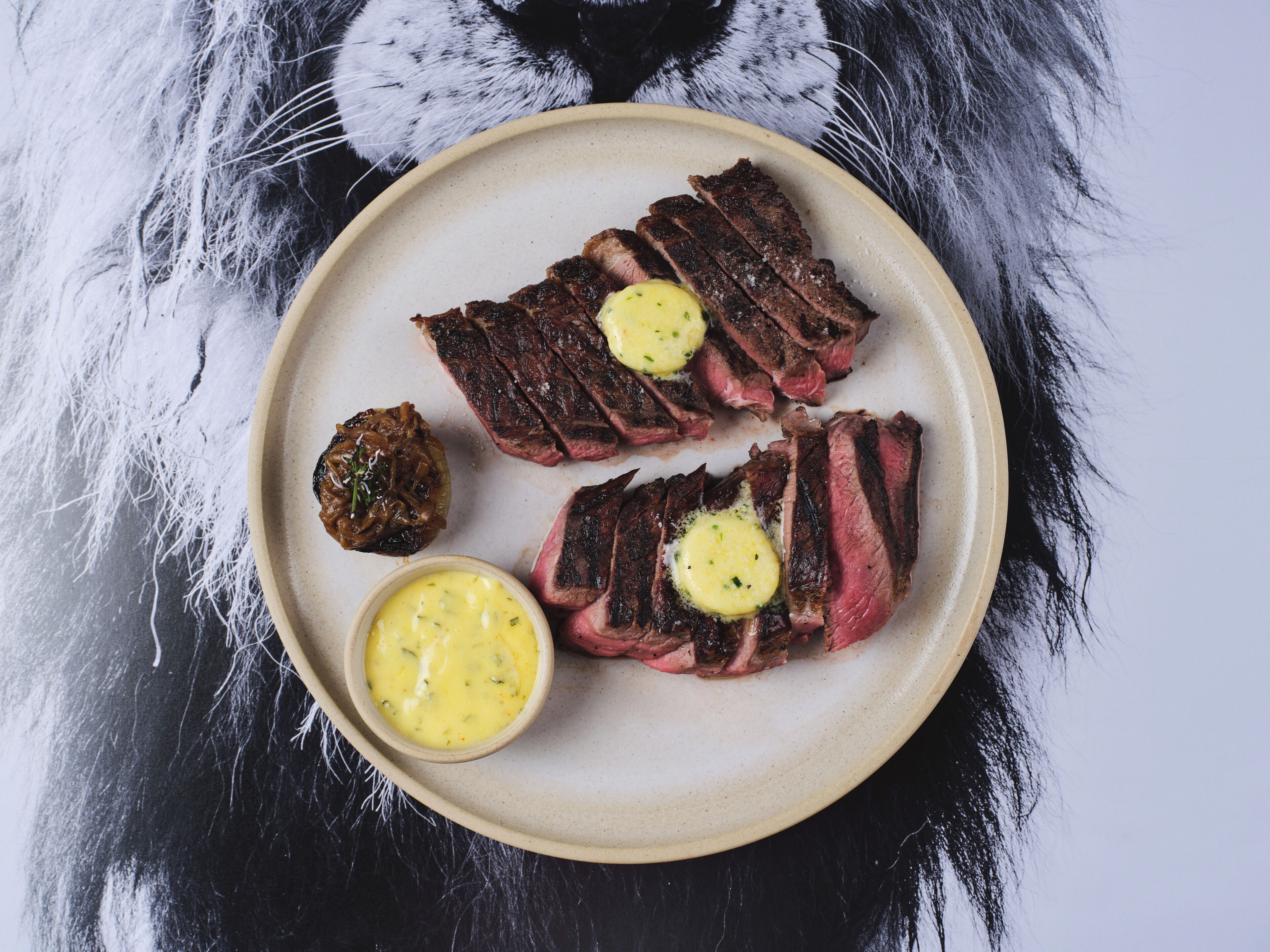 Une assiette contenant un steak et une portion de beurre, posée sur une surface sombre recouverte de fourrure blanche.