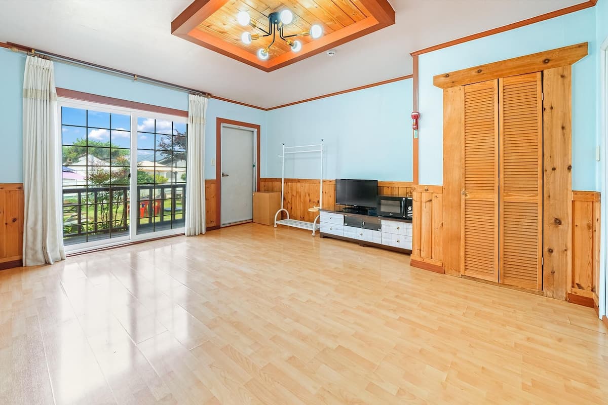 A spacious room with wooden flooring, a sliding glass door to a balcony, a TV, and a wooden wardrobe.