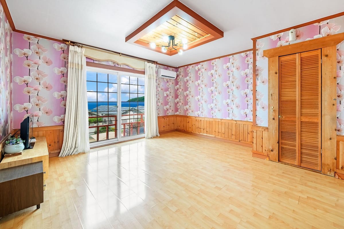 A spacious room with wooden flooring, a large window with curtains, a wooden door, and a ceiling light fixture.