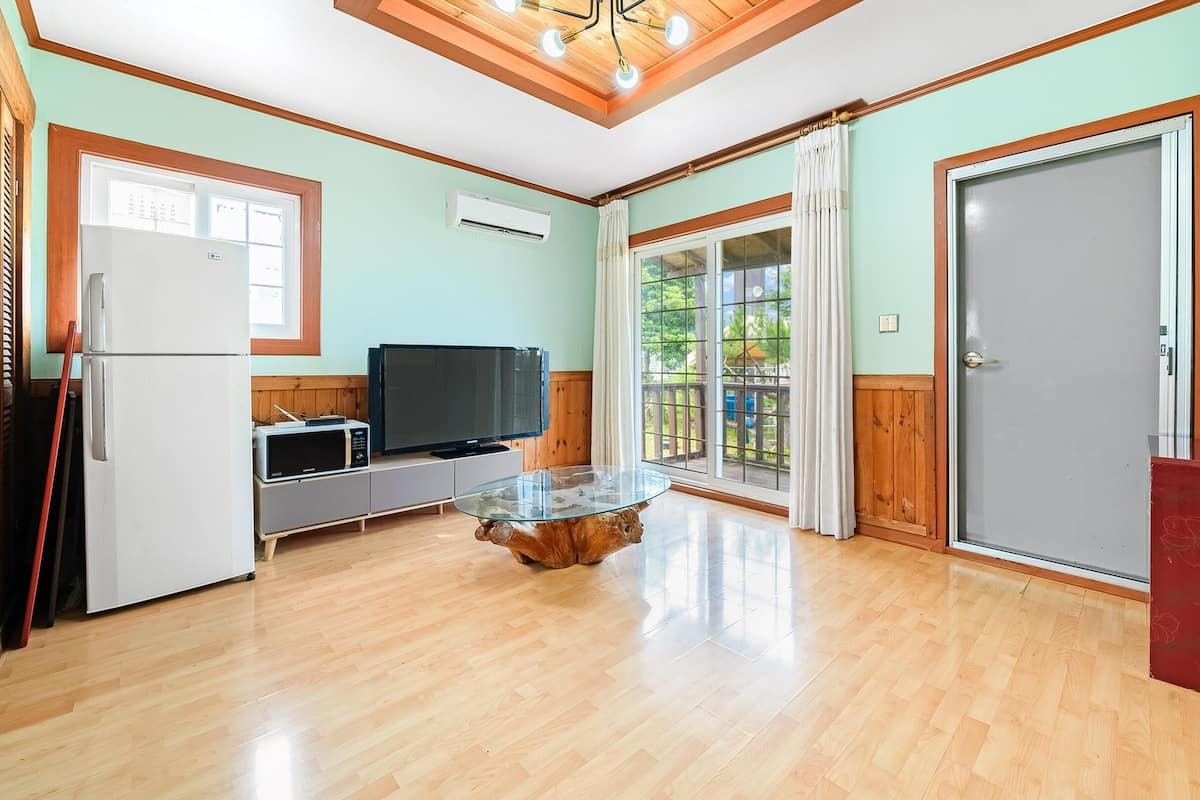 A compact kitchen with a refrigerator, microwave, and TV on a wooden cabinet. There's a glass coffee table in the center, and a sliding glass door leading to a balcony.