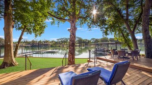 A wooden deck with lounge chairs overlooking a lake, a dock, and a covered seating area with tables and chairs. - NB Lakefront Getaway*Luxury on Lake Dunlap (New Braunfels)
