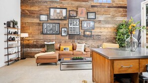 A cozy living room with a wooden wall, a brown leather sofa, a coffee table, and a kitchen area with a wooden cabinet and a vase of flowers. - 8Sages*Texas Forever*Sleeps 6*Private Plunge Pool (New Braunfels)