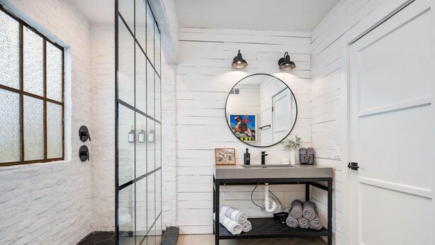 A bathroom with a white brick wall, a glass shower enclosure, a round mirror, a black vanity, and a door. - 8Sages*Texas Forever*Sleeps 6*Private Plunge Pool (New Braunfels)