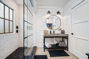 Un baño con pared de ladrillo blanco, cabina de ducha con vidrio, espejo redondo, tocador negro y una puerta.