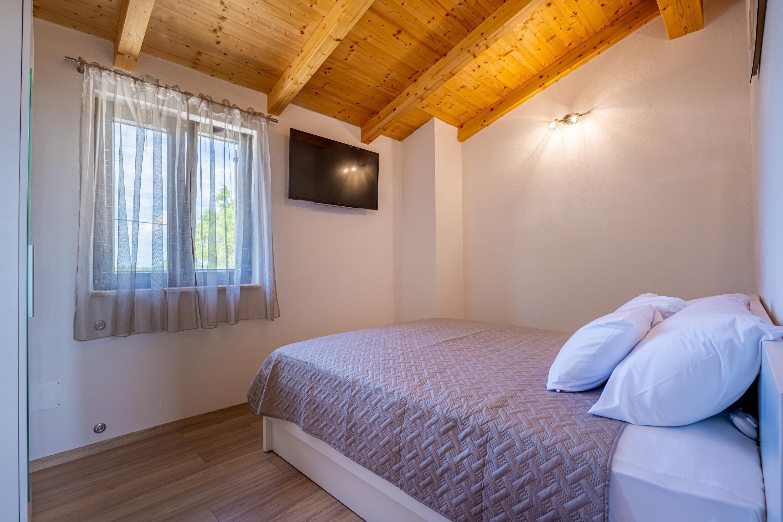 Een slaapkamer met een houten plafond, een bed met witte lakens, een aan de muur bevestigde televisie en een raam met dunne gordijnen.