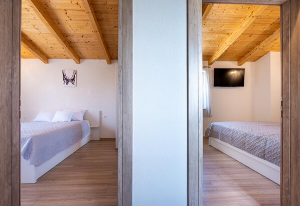 A bedroom with a wooden ceiling, a bed with a white headboard, a wall-mounted TV, and a window with curtains. - Holiday House Balarini (Gajana)