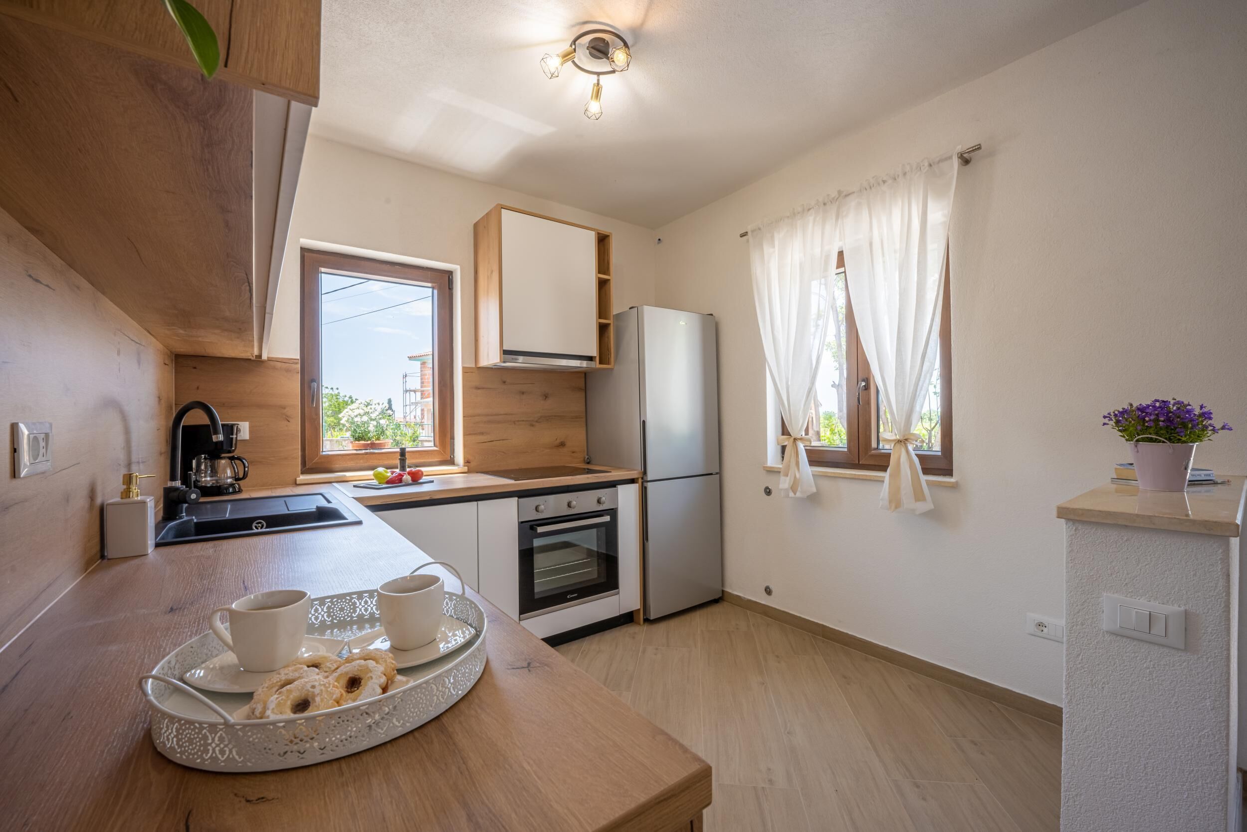 Een moderne keuken met houten kastjes, een roestvrijstalen koelkast, een inbouwoven en een raam met gordijnen.