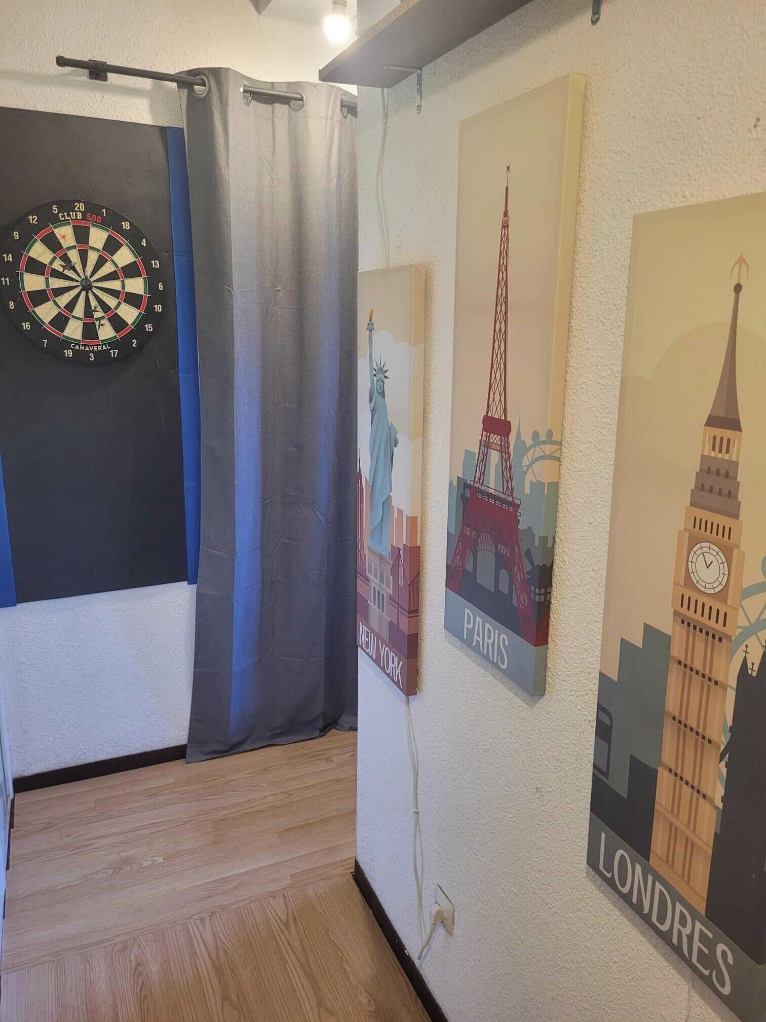 A room with a dartboard, a curtain, and city-themed wall art depicting New York, Paris, and London.
