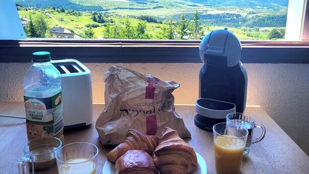 A breakfast setup with croissants, juice, and a bag of pastries on a table by a window with a mountain view. - Appartement Rénové Avec une Magnifique vue (Font-Romeu-Odeillo-Via)