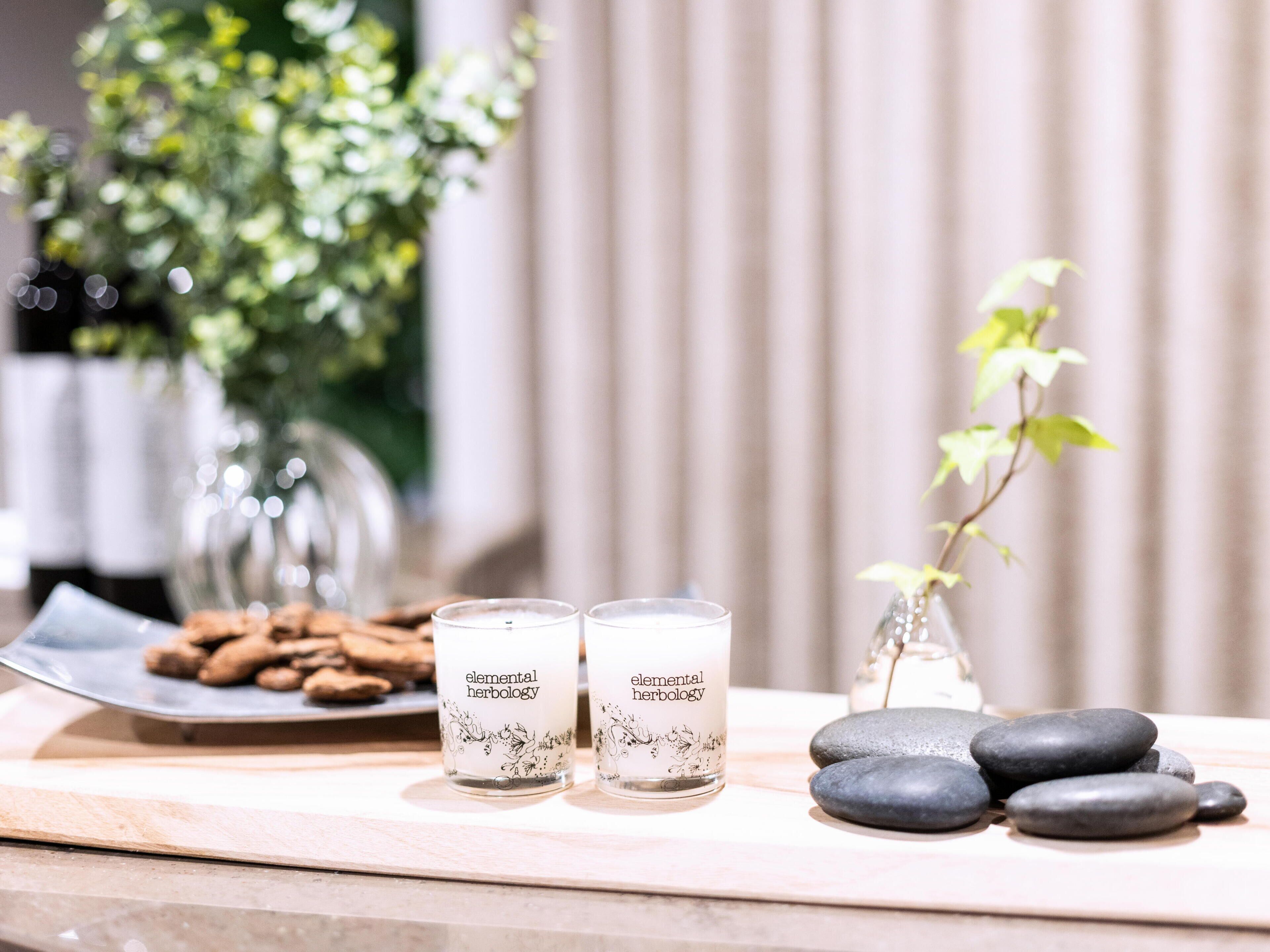 A table with two candles labeled "elemental herbalogy," a plate of cookies, a vase with a plant, and a stack of smooth stones.