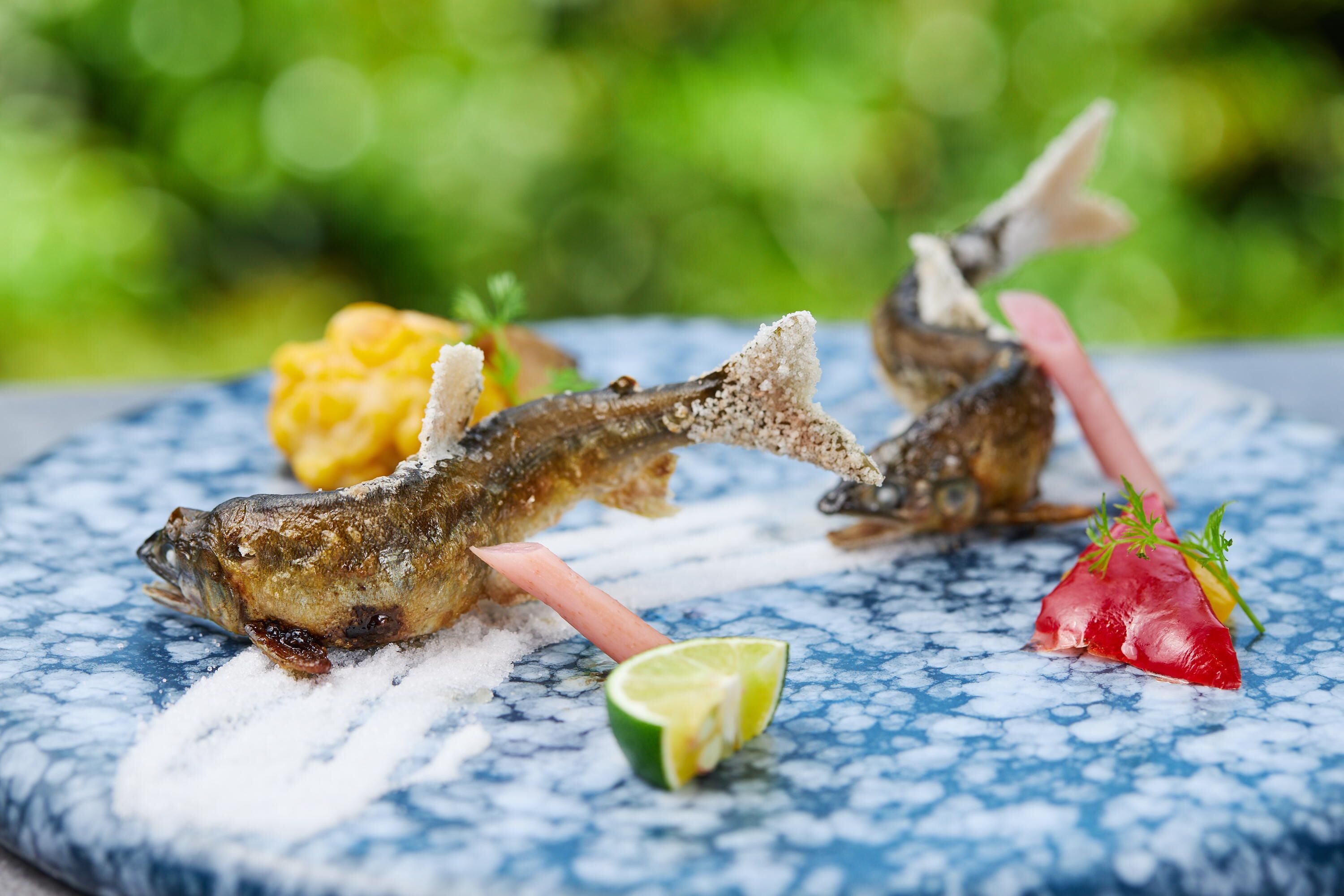 Deux poissons frits servis sur une assiette bleue à motifs, accompagnés d’un quartier de citron vert et d’une garniture.