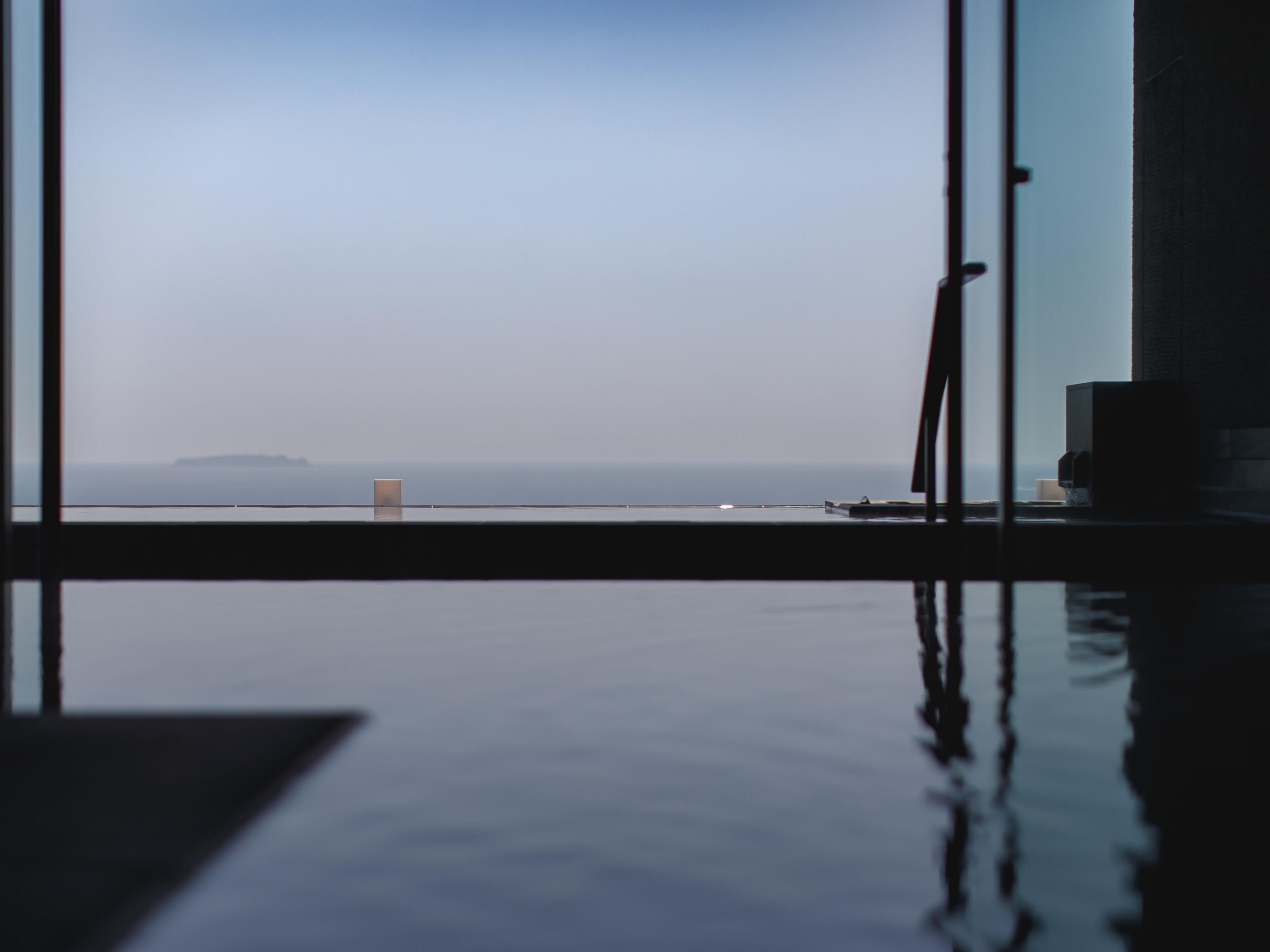 Blick vom Zimmer mit großem Fenster auf ein ruhiges Meer und eine entfernte Insel.