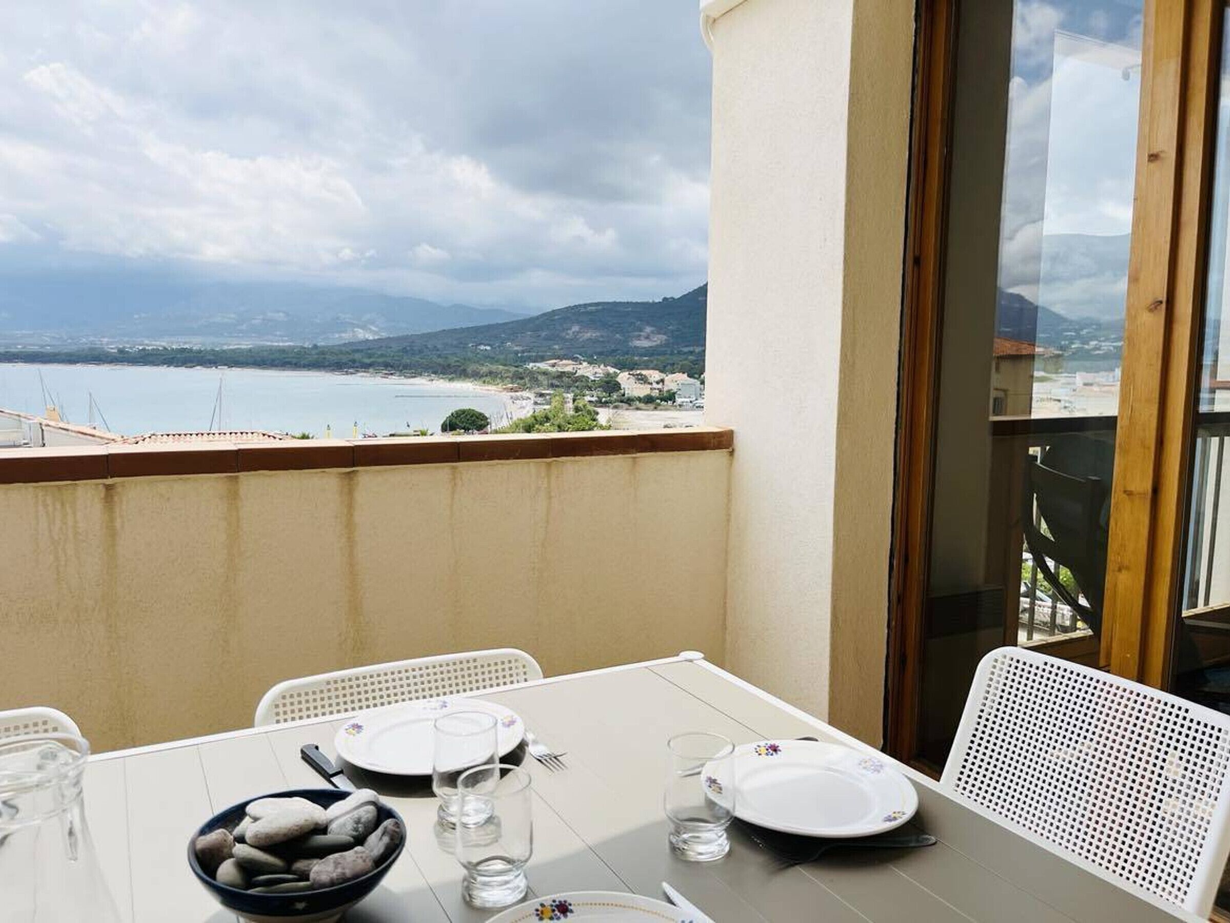 A balcony with a table set for two, overlooking a coastal landscape with mountains in the distance.