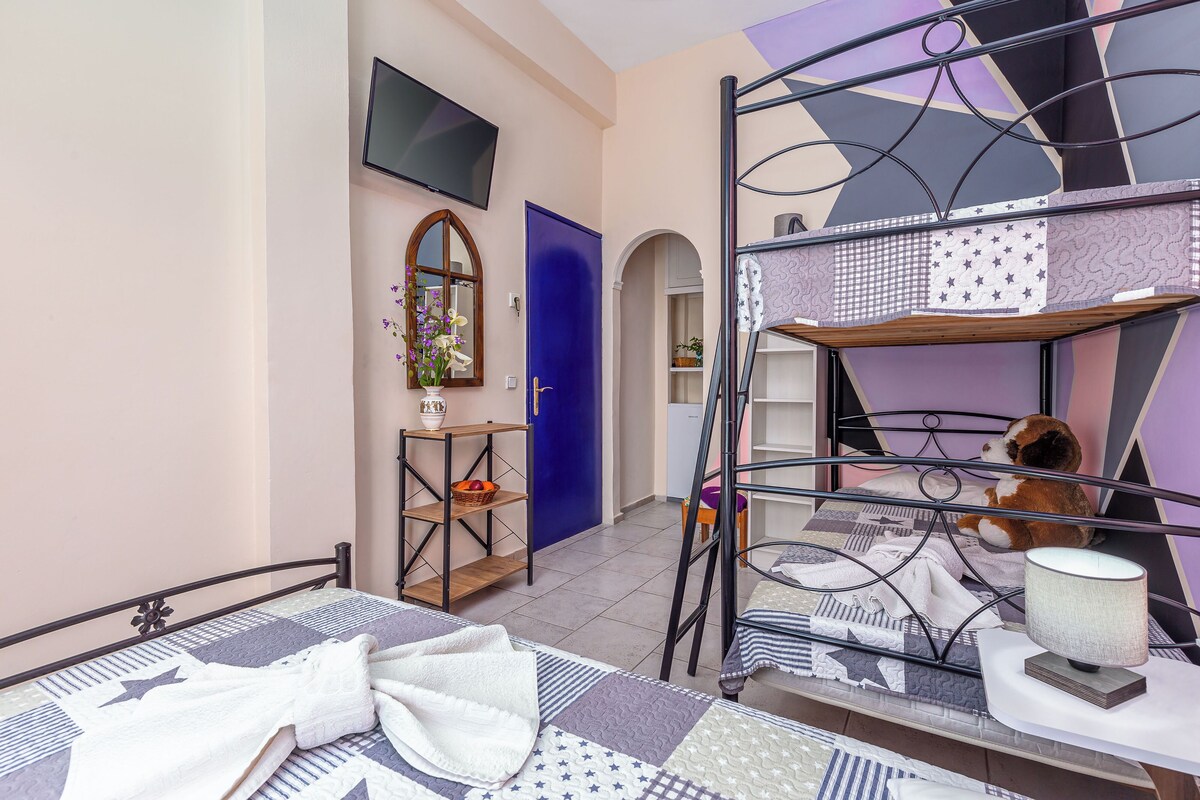 A bedroom with a bunk bed, a TV mounted on the wall, a small table with a lamp, and a door leading to another room.